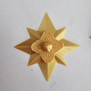 Könnte beinhalten: Goldfarbenes, sternförmiges Dekorationselement mit einem zentralen, vierblättrigen Blumenmuster. Der Stern hat spitze Kanten und eine strukturierte Oberfläche. In der Mitte der Blume befindet sich ein kleiner, goldfarbener Kegel.