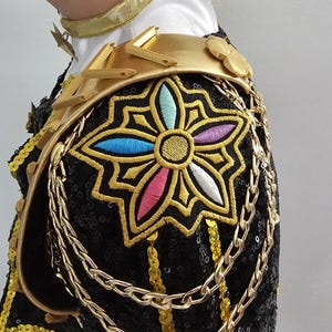 Puede incluir: Primer plano de una prenda negra con lentejuelas y detalles dorados. El hombro presenta una gran flor bordada multicolor, delineada en oro. Cadenas y adornos dorados a&ntilde;aden detalles a esta pieza ornamentada.