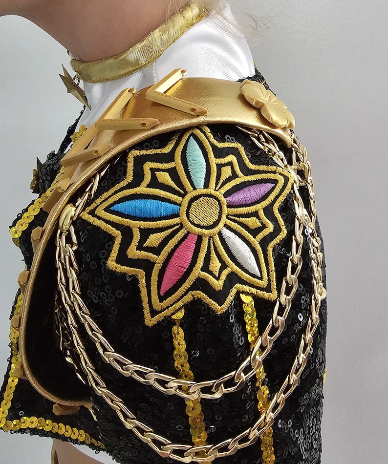 Puede incluir: Primer plano de una prenda negra con lentejuelas y detalles dorados. El hombro presenta una gran flor bordada multicolor, delineada en oro. Cadenas y adornos dorados a&ntilde;aden detalles a esta pieza ornamentada.