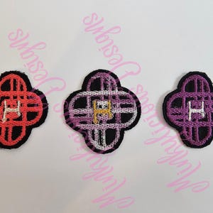 May include: Three embroidered quatrefoil patches. The patches have a black border, with the interior designs in red, purple, and white. Each patch features the letter "H" in the centre.