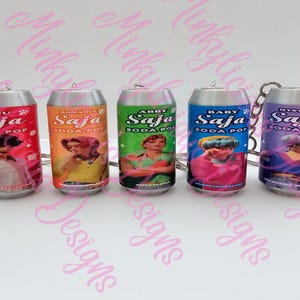 May include: Five miniature soda can keychains. Each can features a different design with the text "Saja Soda Pop" and a unique image. The cans are in shades of red, orange, green, blue, and purple, with silver tops.