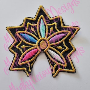 Puede incluir: Parche bordado con dise&ntilde;o de estrella con fondo negro y ribete dorado. La flor central presenta p&eacute;talos rosas, azules y turquesas. El parche es probablemente para ropa o accesorios.