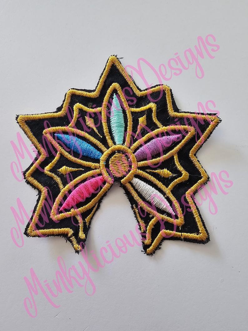 Puede incluir: Parche bordado con dise&ntilde;o de estrella con fondo negro y ribete dorado. La flor central presenta p&eacute;talos rosas, azules y turquesas. El parche es probablemente para ropa o accesorios.