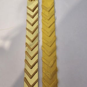 以下が含まれることがあります： 2つの金色の長方形の装飾品。1つは、金色の背景に茶色の山形模様が並んでいます。もう1つは金色の山形模様です。どちらも上部に金の花が付いています。工芸品や装飾用である可能性があります。