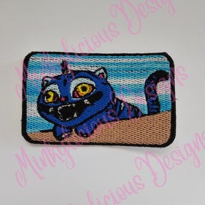 May include: Embroidered patch featuring a blue cartoon cat with yellow eyes and a black mouth. The cat is set against a blue and white striped background, with a brown border. The patch has a rectangular shape.
