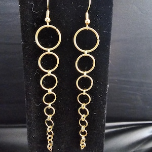 May include: A pair of gold-colored dangle earrings. Each earring features a series of connected gold rings of varying sizes, culminating in a small chain at the bottom. The earrings are displayed against a black background.