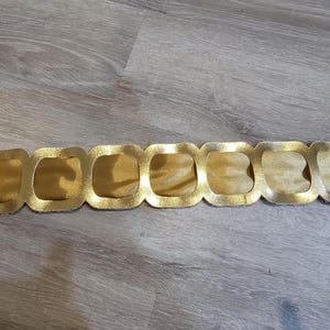 Op de afbeelding: Een goudkleurige riem met een reeks vierkante gespen met een open midden. Het materiaal van de riem lijkt een zachte, flexibele stof te zijn. De gespen zijn glanzend goudkleurig en de riem ligt op een houten oppervlak.