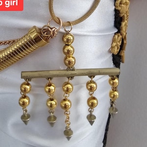 May include: Close-up of a gold-toned necklace with multiple decorative elements. The necklace features a series of gold beads and dangling cone-shaped pendants. A textured gold tube and a gold chain are also visible. The background is a white fabric.