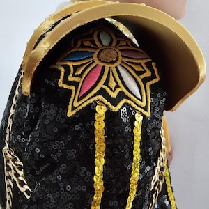 Puede incluir: Una hombrera negra con lentejuelas con adornos y adornos dorados. La hombrera presenta un cuello dorado y un dise&ntilde;o floral bordado con acentos rosas, azules y blancos. Cadenas doradas y lentejuelas colgantes a&ntilde;aden detalles.