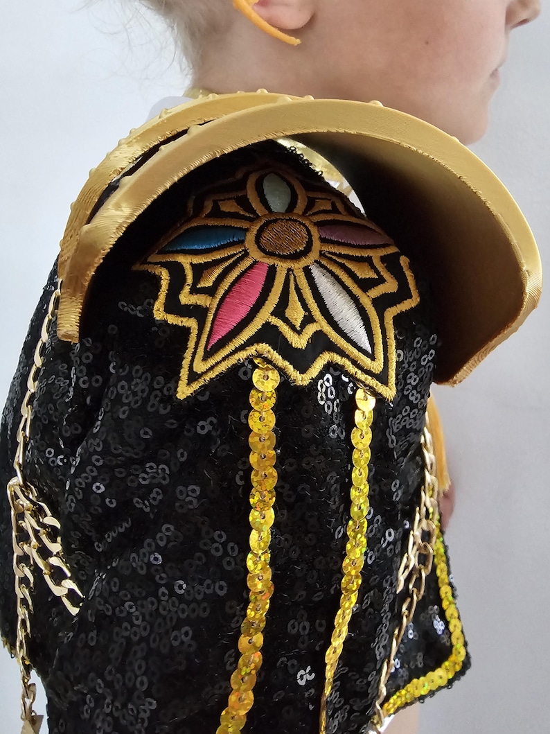 Puede incluir: Una hombrera negra con lentejuelas con adornos y adornos dorados. La hombrera presenta un cuello dorado y un dise&ntilde;o floral bordado con acentos rosas, azules y blancos. Cadenas doradas y lentejuelas colgantes a&ntilde;aden detalles.