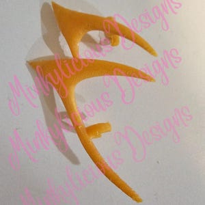 May include: Two orange-yellow, pointed ear-shaped accessories, possibly cosplay or costume pieces, are displayed against a white background. The accessories have a curved design with a small tab on the side. The image has the text "Minklycious Designs" in a pink cursive font.