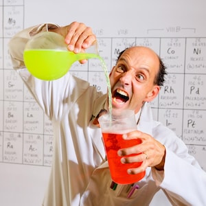 May include: A scientist in a white lab coat pours neon green liquid from a flask into a glass of red liquid. The background features a periodic table of elements.