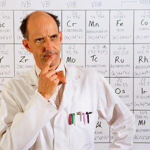 May include: A man in a white lab coat stands in front of a periodic table of elements, deep in thought. He wears a bow tie and has pens in his pocket. The background is a white board with handwritten elements.
