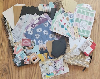 May include: A collection of craft supplies for scrapbooking and journaling, including patterned paper, stickers, washi tape, and decorative embellishments. The supplies are arranged on a wooden surface.