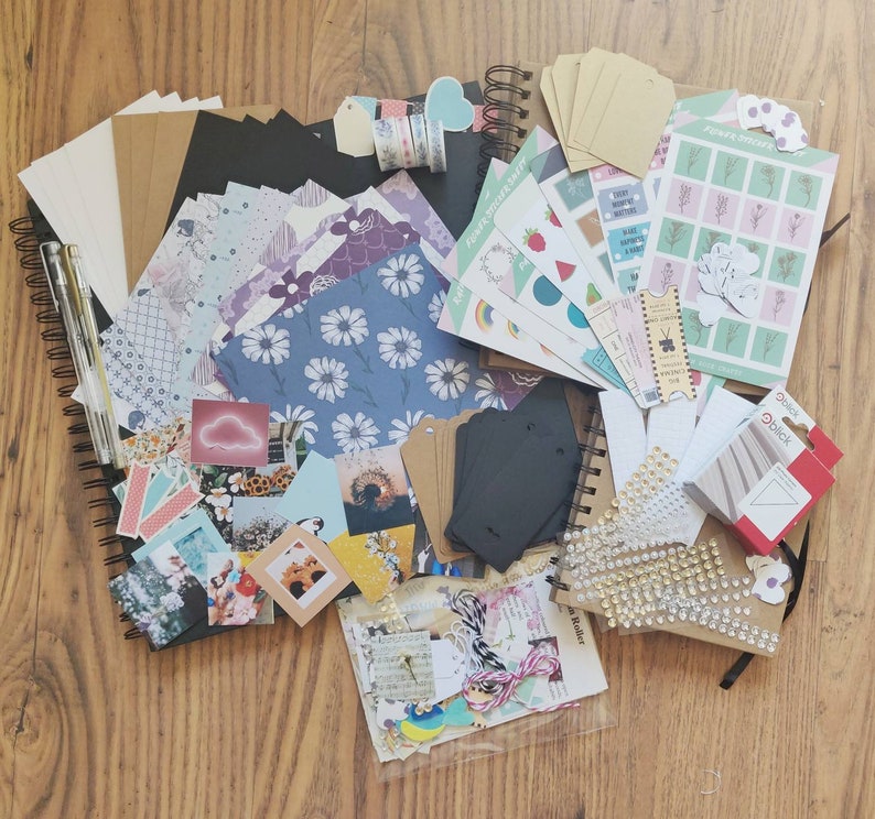 K&ouml;nnte beinhalten: Eine Sammlung von Bastelartikeln zum Scrapbooking und Journaling, darunter gemustertes Papier, Aufkleber, Washi-Tape und dekorative Verzierungen. Die Materialien sind auf einer Holzoberfl&auml;che angeordnet.