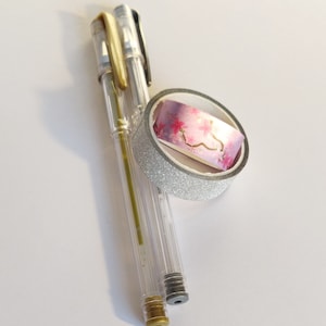 May include: Two clear gel pens with gold and black accents positioned next to a roll of decorative washi tape. The tape features a silver glitter exterior and an inner pattern of pink cherry blossoms with gold foil branches on a gradient background.
