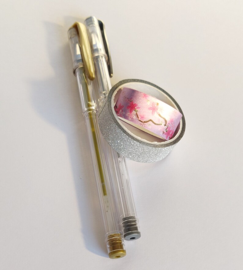May include: Two clear gel pens with gold and black accents positioned next to a roll of decorative washi tape. The tape features a silver glitter exterior and an inner pattern of pink cherry blossoms with gold foil branches on a gradient background.