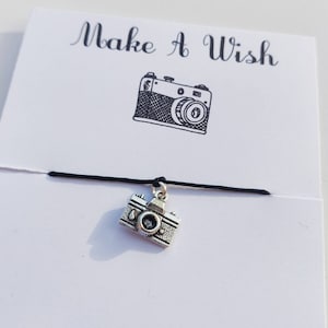 Peut inclure: Un bracelet en cordon noir avec un charme en argent en forme d'appareil photo. Le charme est détaillé et possède un petit objectif. Le bracelet est sur une carte blanche avec le texte "Make a Wish" imprimé dessus.