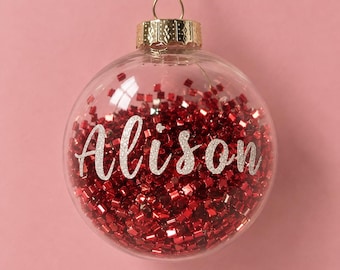May include: A clear spherical Christmas ornament filled with small, shiny red square-shaped confetti. The name Alison is written across the center in white glittery cursive script. The ornament has a gold-colored metal cap and a gold hanging loop.