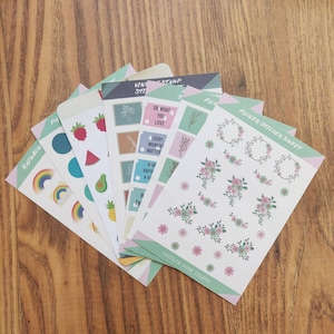 K&ouml;nnte beinhalten: Ein Set mit sechs Stickerb&ouml;gen mit verschiedenen Designs. Die B&ouml;gen enthalten einen Regenbogen, Fr&uuml;chte, Blumen und Briefmarken mit motivierenden Zitaten. Die B&ouml;gen sind auf wei&szlig;em Papier mit einem hellgr&uuml;nen Rand gedruckt.