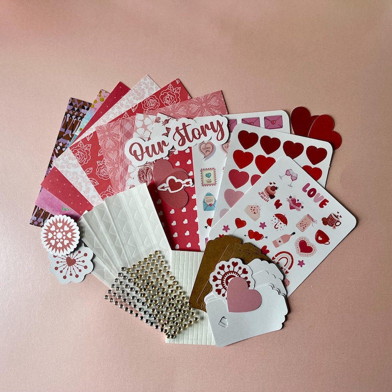 May include: A collection of craft supplies including patterned paper sheets in shades of red and pink, a sticker sheet with heart and love-themed icons, a die-cut sign reading Our Story, white and brown gift tags, and a sheet of small adhesive gems.