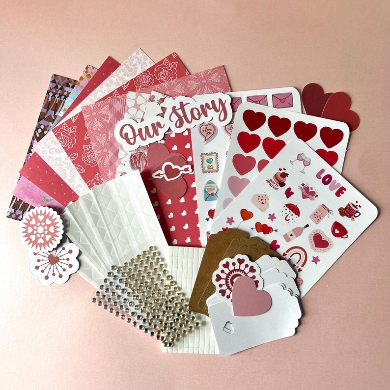May include: A collection of craft supplies featuring patterned paper sheets in shades of red and pink, sticker sheets with heart and love-themed icons, a sheet of adhesive rhinestones, and several blank white and brown cardstock gift tags.