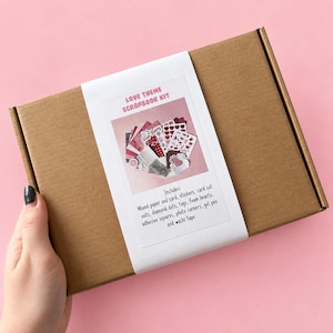 May include: A brown cardboard box featuring a white label with the text LOVE THEME SCRAPBOOK KIT. The label displays an image of various red and white paper, stickers, and tags, with a list of contents including paper, card, stickers, and tape.