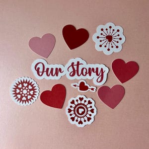 May include: A collection of paper cutouts featuring the text Our Story in red script, surrounded by various red and pink heart shapes, circular floral-like patterns with heart motifs, and a small heart pierced by an arrow on a light background.