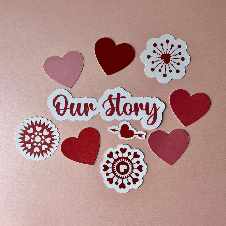 May include: A collection of paper cutouts featuring the text Our Story in red script, surrounded by various red and pink heart shapes, circular floral-like patterns with heart motifs, and a small heart pierced by an arrow on a light background.