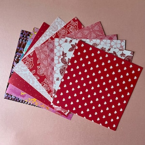 May include: A collection of square paper sheets arranged in a fan shape. The patterns include red and white hearts, various floral designs, geometric shapes, and abstract prints in shades of red, pink, white, purple, and gold foil accents.