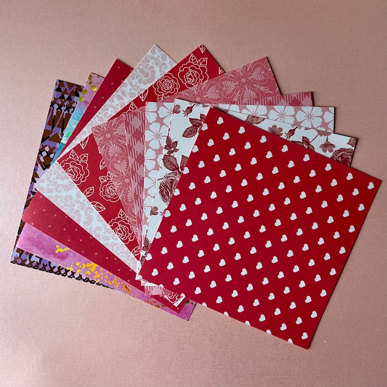 May include: A collection of square paper sheets arranged in a fan shape. The patterns include red and white hearts, various floral designs, geometric shapes, and abstract prints in shades of red, pink, white, purple, and gold foil accents.