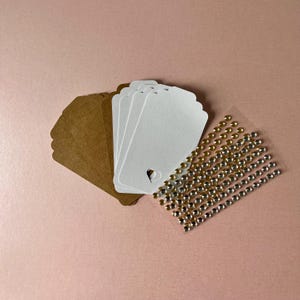 May include: A collection of rectangular paper tags in brown and white, featuring a small heart-shaped cutout at the bottom of each white tag. Beside the tags is a clear adhesive sheet containing rows of small, round, metallic gold and silver stickers.