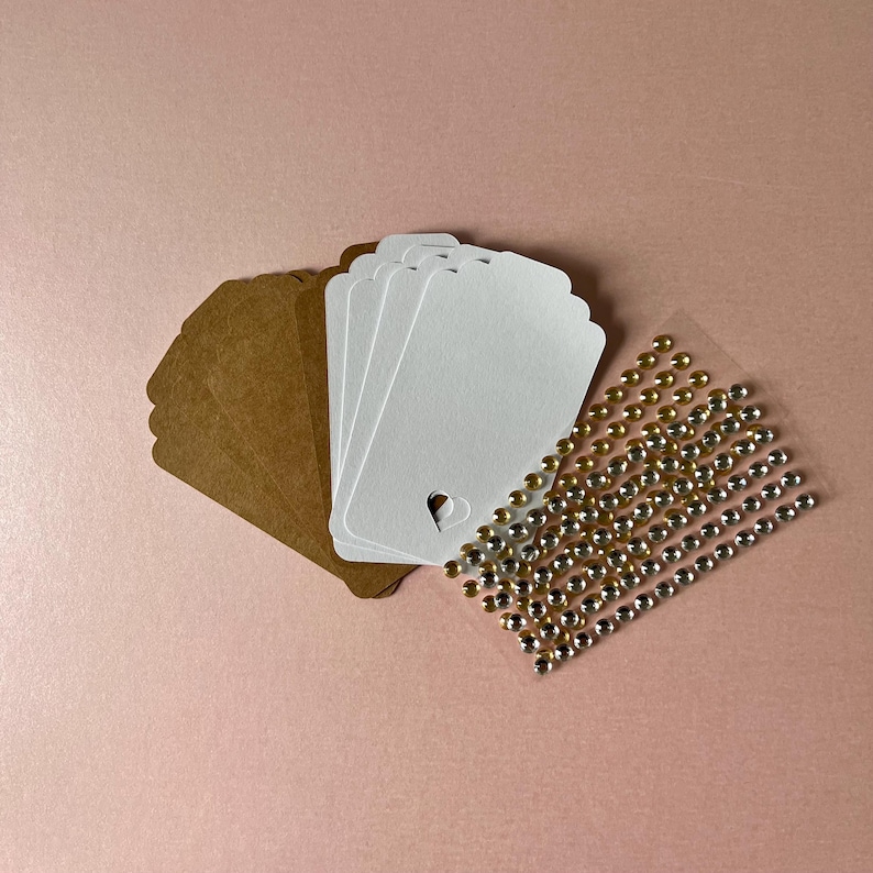 May include: A collection of rectangular paper tags in brown and white, featuring a small heart-shaped cutout at the bottom of each white tag. Beside the tags is a clear adhesive sheet containing rows of small, round, metallic gold and silver stickers.