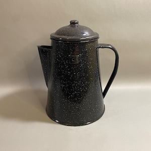 May include: A black enamel coffee pot with a speckled pattern. The pot has a lid, a spout, and a handle. The pot is made of metal and has a vintage look. The coffee pot is a classic kitchenware item.