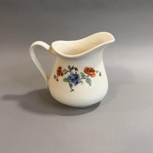 May include: Cream-colored ceramic creamer with a curved spout and handle. The creamer is decorated with a floral pattern in shades of orange, blue, and green. The creamer is a classic design for serving cream or milk.