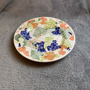 May include: A white ceramic plate with a grapevine design. The plate features green leaves, brown vines, and bunches of purple grapes.