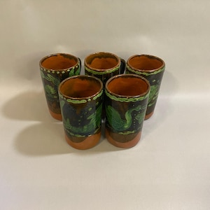 Puede incluir: Un juego de cinco vasos de chupito de cer&aacute;mica hechos a mano. Cada vaso presenta un exterior marr&oacute;n oscuro con detalles verdes y naranjas. Los vasos tienen forma cil&iacute;ndrica y est&aacute;n decorados con un dise&ntilde;o de hojas verdes.