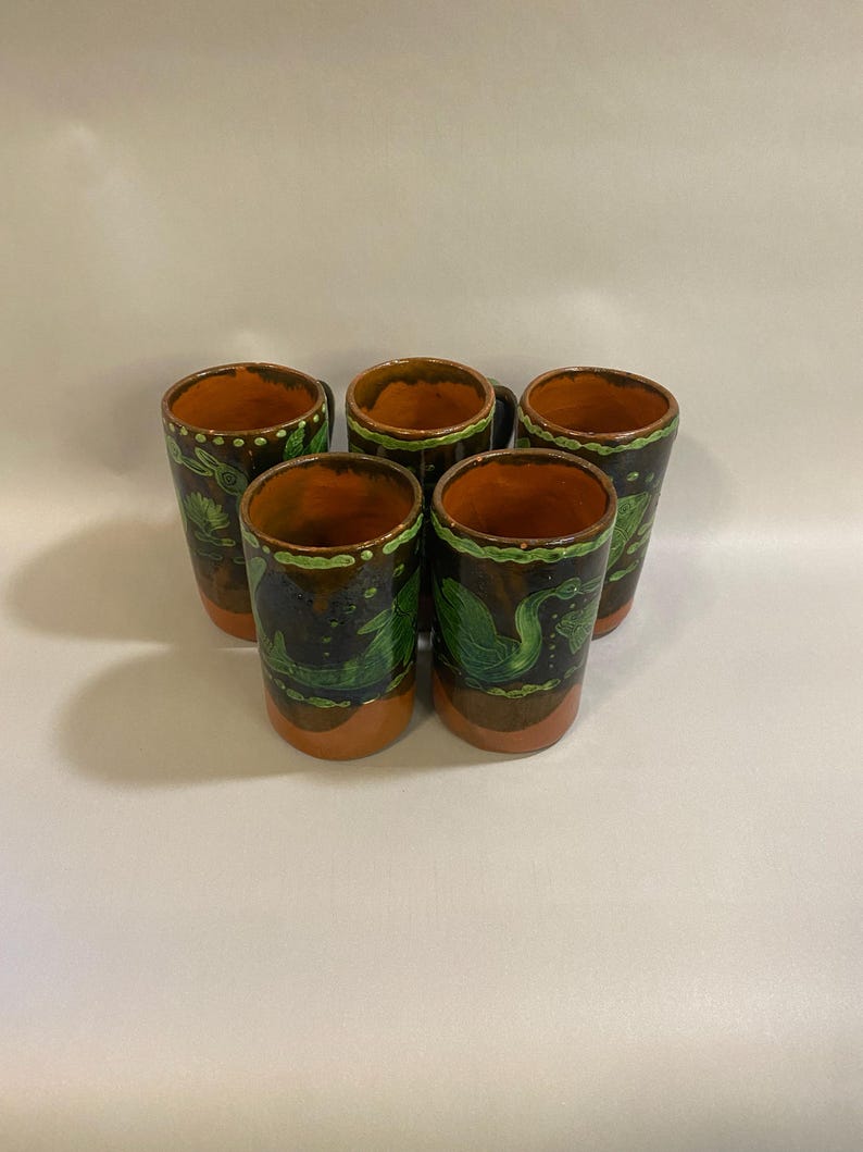 Puede incluir: Un juego de cinco vasos de chupito de cer&aacute;mica hechos a mano. Cada vaso presenta un exterior marr&oacute;n oscuro con detalles verdes y naranjas. Los vasos tienen forma cil&iacute;ndrica y est&aacute;n decorados con un dise&ntilde;o de hojas verdes.