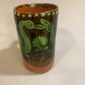 Puede incluir: Vaso de cer&aacute;mica hecho a mano con exterior marr&oacute;n e interior naranja. El vaso presenta dise&ntilde;os pintados en verde de aves y un nen&uacute;far. El borde est&aacute; punteado con pintura verde. La base es naranja.