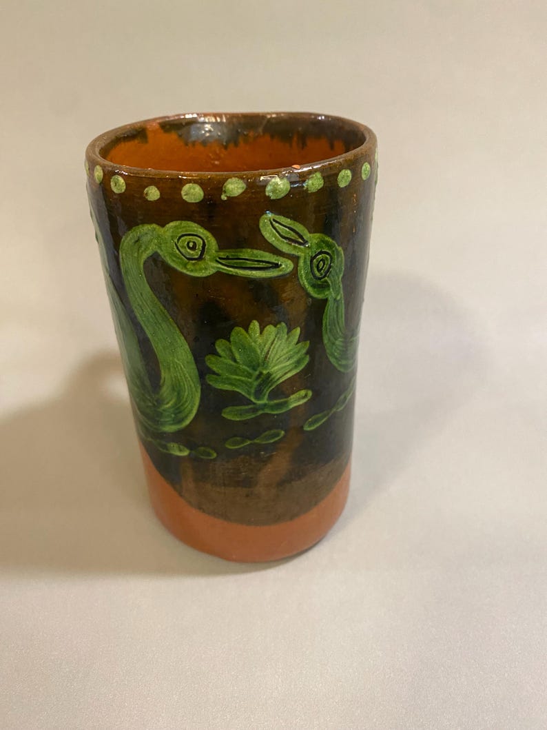Puede incluir: Vaso de cer&aacute;mica hecho a mano con exterior marr&oacute;n e interior naranja. El vaso presenta dise&ntilde;os pintados en verde de aves y un nen&uacute;far. El borde est&aacute; punteado con pintura verde. La base es naranja.