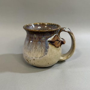 May include: Handmade ceramic mug with a unique leaf-shaped handle attachment. The mug features a blend of colors, including brown, beige, and blue, creating a rustic and artistic appearance. This handcrafted mug is perfect for enjoying coffee or tea.