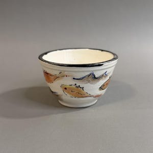 May include: A small, off-white ceramic bowl with a black rim. The bowl is decorated with hand-painted orange, yellow, and blue fish. The bowl is a decorative piece for serving snacks or small portions.