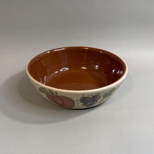 May include: A ceramic bowl with a brown interior and a cream-colored exterior. The exterior features a painted design of fruit, including apples and grapes. The bowl is round and has a wide rim.