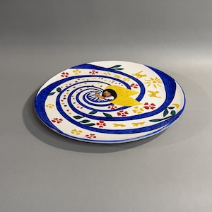 May include: A white ceramic plate with a blue spiral design. The plate features a central image of a person with yellow wings. The design includes red and yellow flowers, green leaves, and yellow birds.