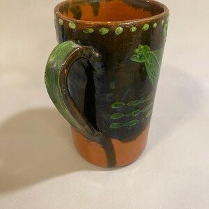 Puede incluir: Taza de cer&aacute;mica hecha a mano con un esmalte marr&oacute;n oscuro, acentuada con detalles en naranja y verde. La taza presenta un asa verde y patrones decorativos de hojas y puntos verdes. Una pieza &uacute;nica y r&uacute;stica.
