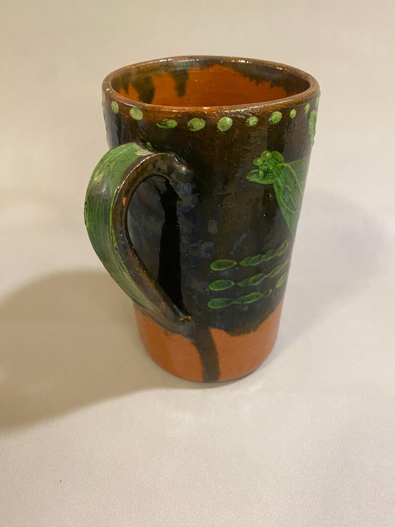 Puede incluir: Taza de cer&aacute;mica hecha a mano con un esmalte marr&oacute;n oscuro, acentuada con detalles en naranja y verde. La taza presenta un asa verde y patrones decorativos de hojas y puntos verdes. Una pieza &uacute;nica y r&uacute;stica.