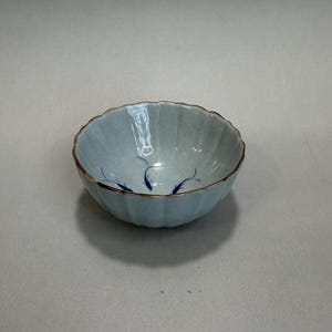 May include: A small, light blue ceramic bowl with a scalloped rim and a gold-colored edge. The bowl features a subtle, ribbed texture and is decorated with blue fish motifs. This decorative bowl is ideal for serving small portions.