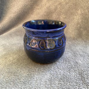 May include: A small, blue ceramic pot with a textured, hand-shaped design. The pot has a dark blue glaze and a brown rim.