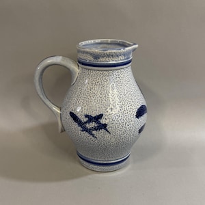 May include: A ceramic pitcher with a speckled white and blue design. The pitcher has a curved handle and a spout. It is decorated with blue floral motifs and blue bands around the top and bottom.