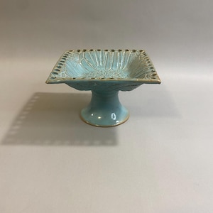 May include: A turquoise ceramic pedestal bowl with a square top and a round base. The bowl features a detailed floral pattern on the inside and a decorative border. The pedestal bowl is a decorative item.